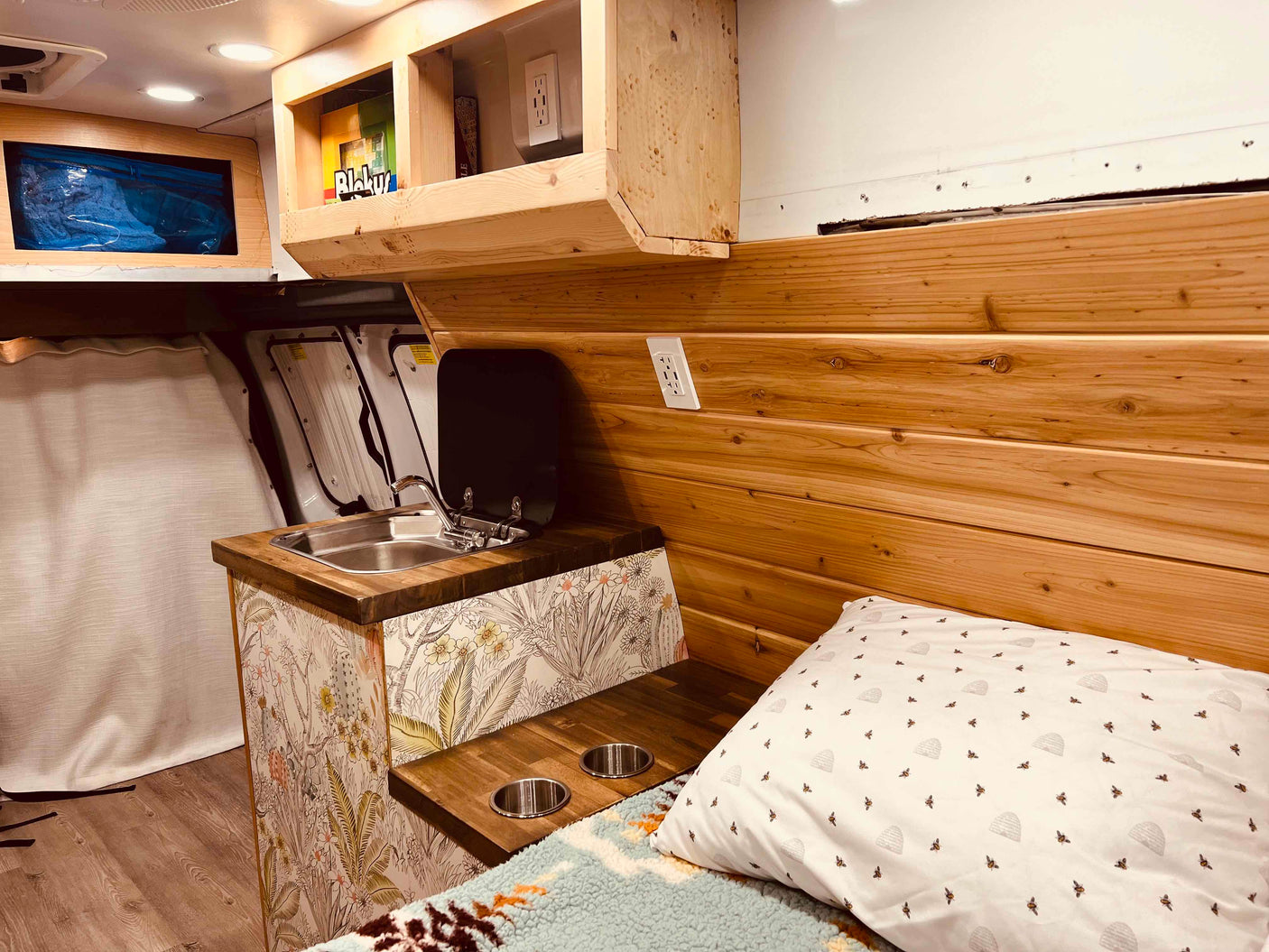 Bright rv rental interior: wood-paneled walls, mini kitchen with sink, patterned cabinet, cozy bed, and storage.