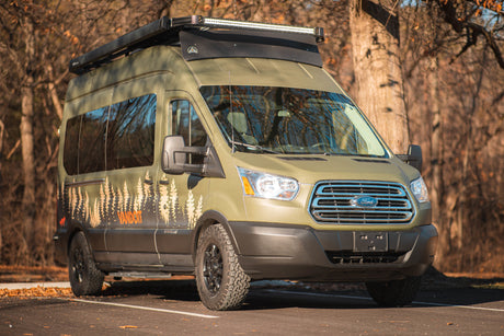 An olive green Ford Transit camper van with custom tree graphics, off-road tires, and a roof rack, ready for your next RV rental trip.