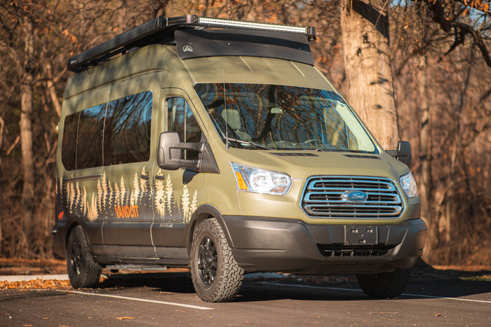 An olive green Ford Transit camper van with custom tree graphics, off-road tires, and a roof rack, ready for your next RV rental trip.