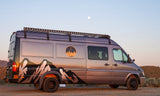 This grey camper van, ideal for RV rental adventures, features mountain graphics and a roof rack under a soft, moonlit sky.