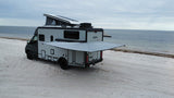 Adventure awaits! This modern RV rental, with its pop-top open & awning extended, sits peacefully on a white sand beach.