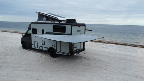 Adventure awaits! This modern RV rental, with its pop-top open & awning extended, sits peacefully on a white sand beach.