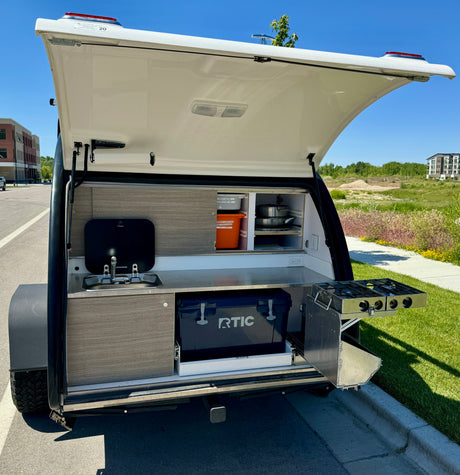 This RV rental teardrop trailer features a compact outdoor kitchen with a sink, two-burner stove, and RTIC cooler, ideal for camping.