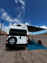 Enjoying an RV rental adventure! Rear view of a white camper van with an extended awning, table, and chairs at a scenic campsite setup.