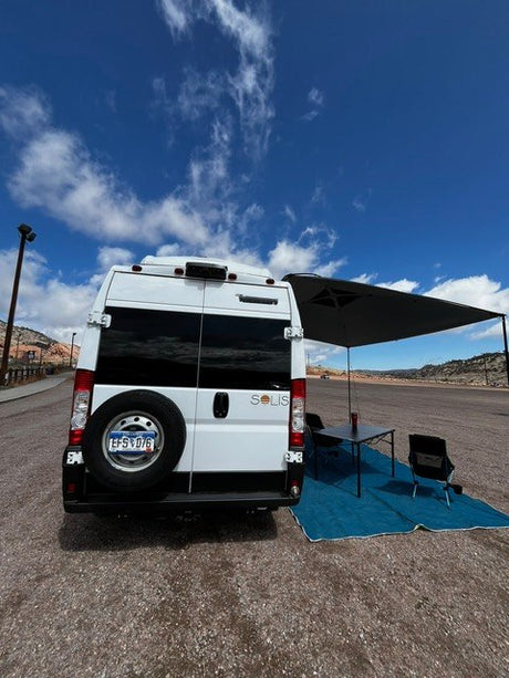 Enjoying an RV rental adventure! Rear view of a white camper van with an extended awning, table, and chairs at a scenic campsite setup.