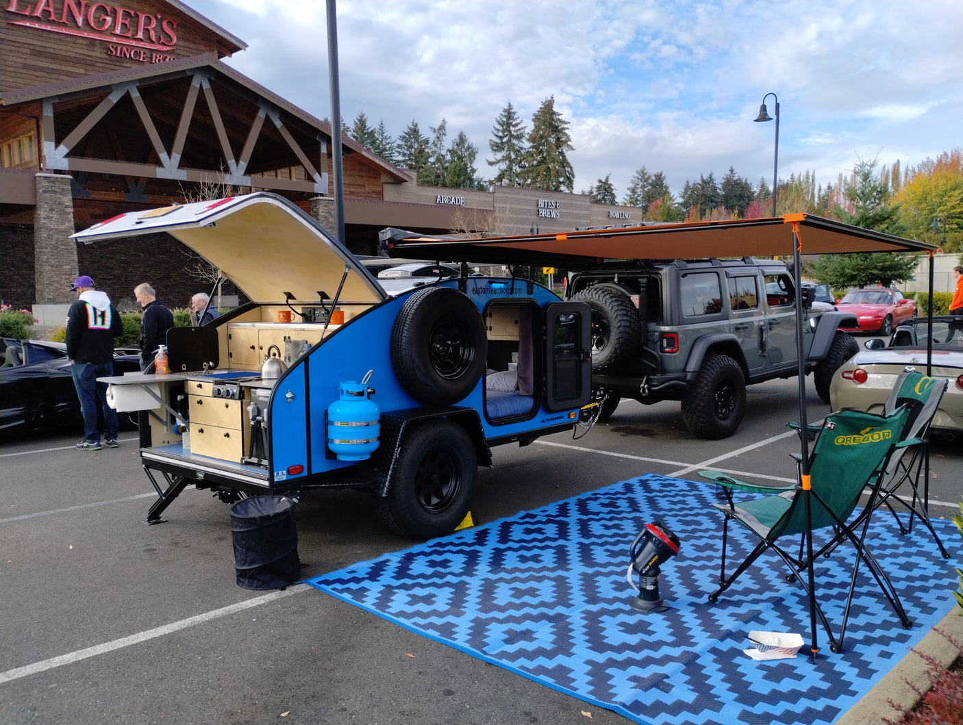 Compact blue teardrop trailer RV rental displayed with Jeep & camping gear. Ideal for outdoor adventures, set up in a parking lot.