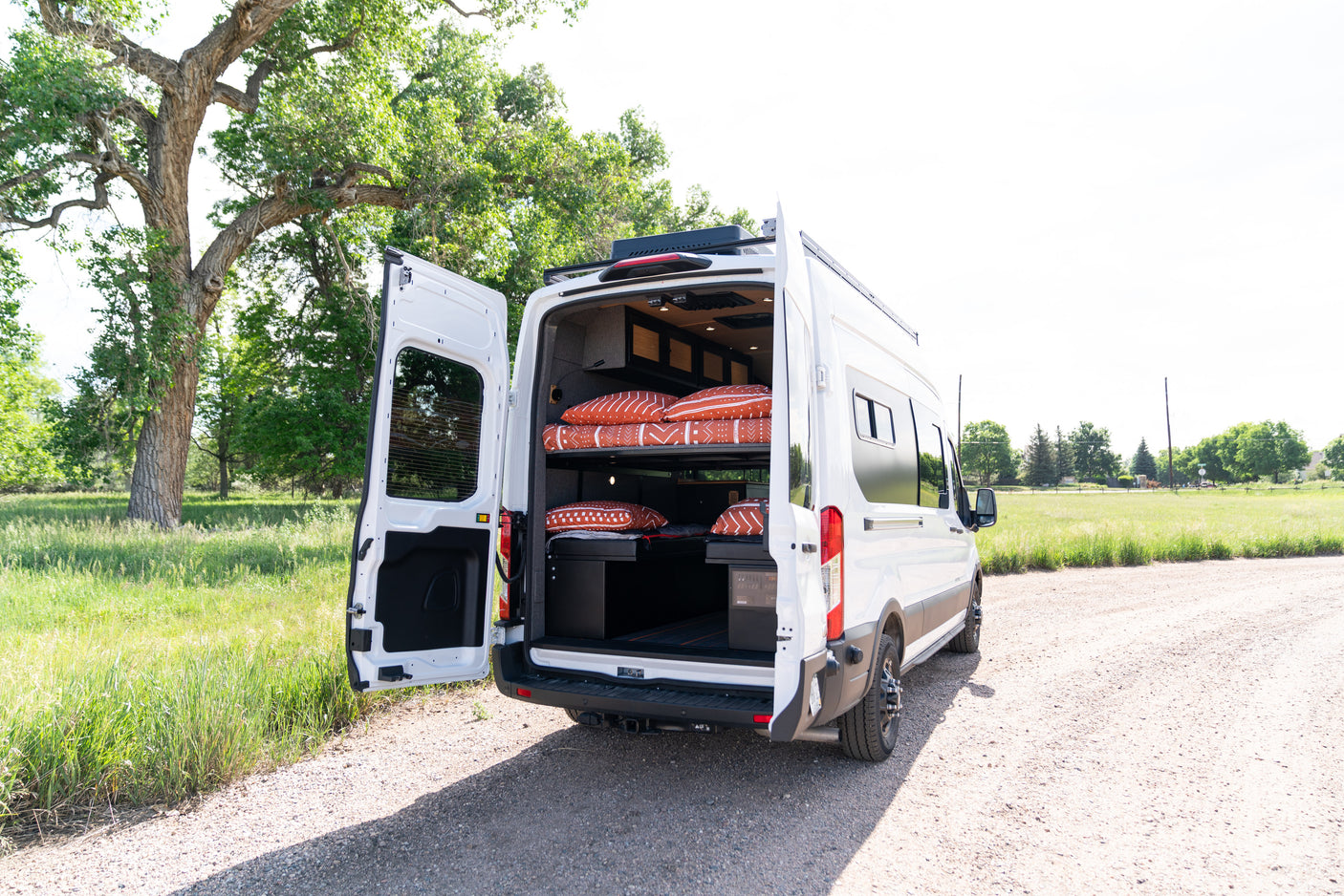 White RV rental camper van with open back doors reveals stacked bunk beds, parked on a dirt road amidst green trees.