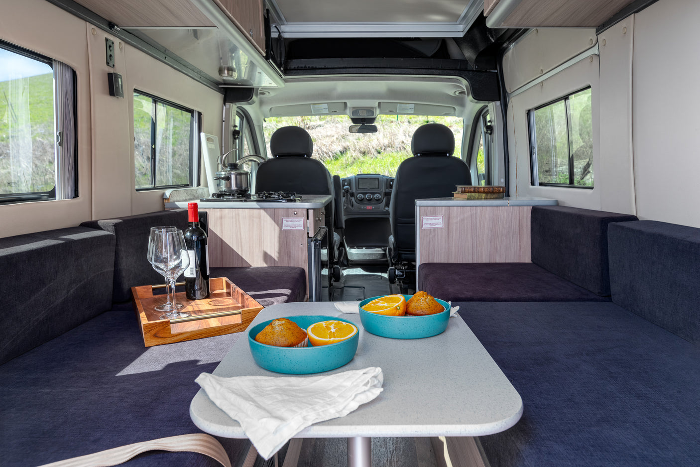 Inviting rv rental interior with dining area, compact kitchen, comfy seating, and large windows revealing green outdoor views.