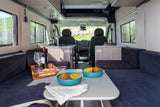 Inviting rv rental interior with dining area, compact kitchen, comfy seating, and large windows revealing green outdoor views.