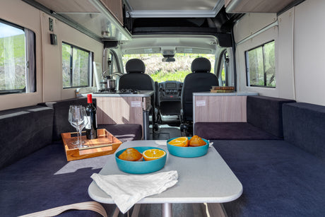 Inviting rv rental interior with dining area, compact kitchen, comfy seating, and large windows revealing green outdoor views.