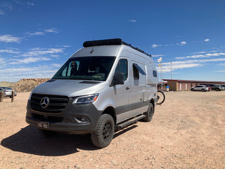 Silver Mercedes Sprinter van, an ideal RV rental, in a dusty lot. Dog in window, ready for adventure!