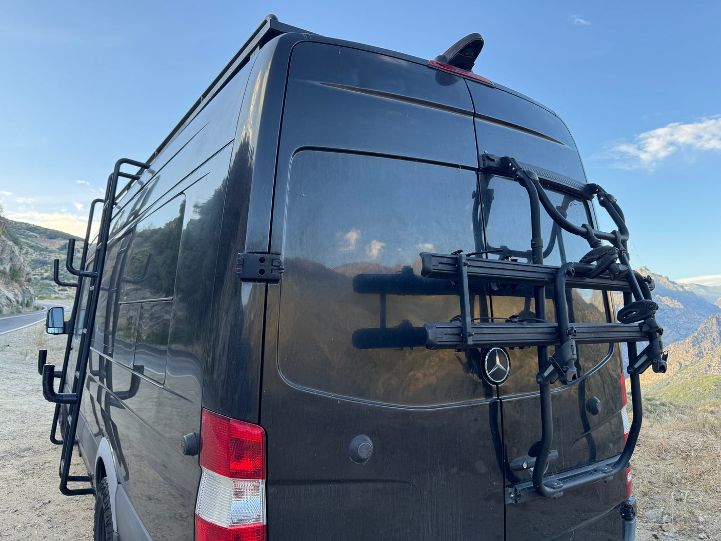 Dark Mercedes-Benz Sprinter RV rental with rear bike rack and side ladder, ready for adventure in the mountains.