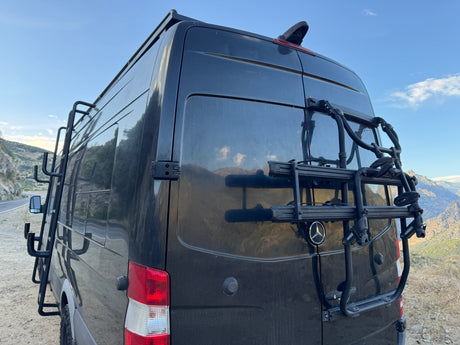 Dark Mercedes-Benz Sprinter RV rental with rear bike rack and side ladder, ready for adventure in the mountains.