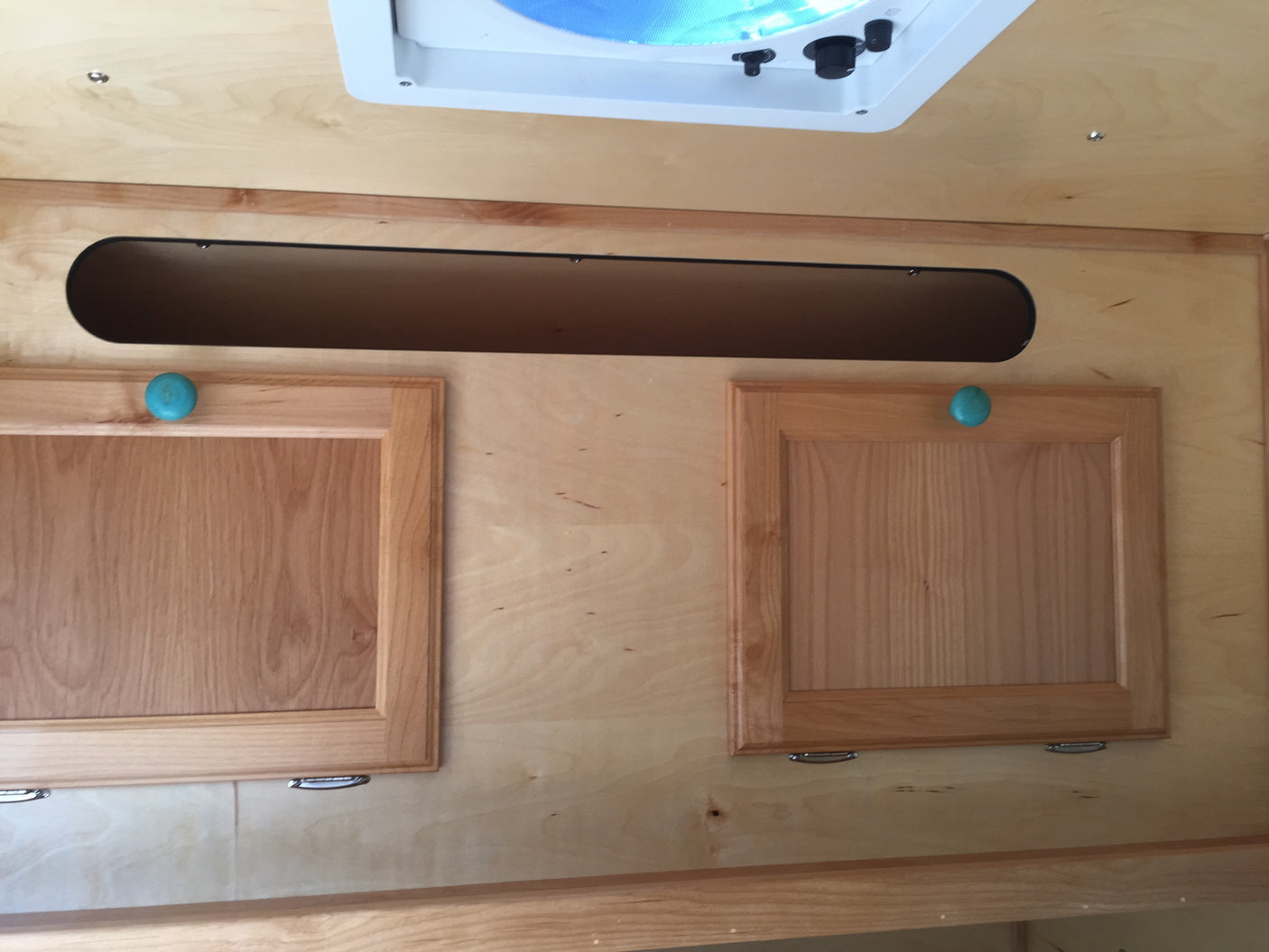 RV rental interior showcasing light wood paneling, cabinets with teal knobs, and an overhead storage cubby with a ceiling vent.