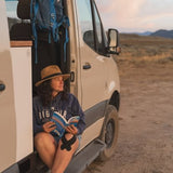 Woman in a hat with athletic tape on her knee reads a book in her RV rental. Parked in open nature at sunset.