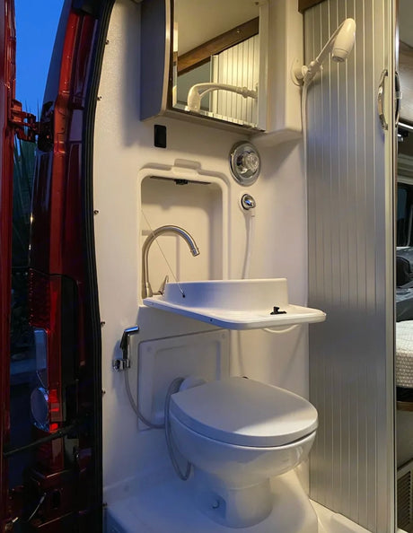 Interior of an RV rental showing a white, compact wet bath with toilet, sink, shower, and mirrored cabinet.
