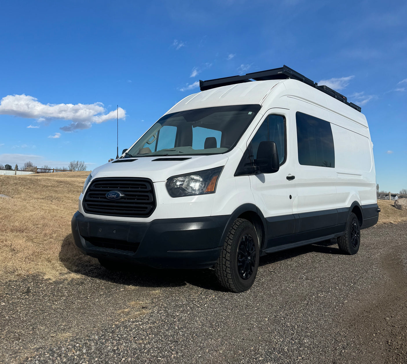 White Ford Transit campervan, ideal for your next RV rental adventure. Parked on a gravel lot under a clear blue sky.