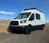 White Ford Transit campervan, ideal for your next RV rental adventure. Parked on a gravel lot under a clear blue sky.