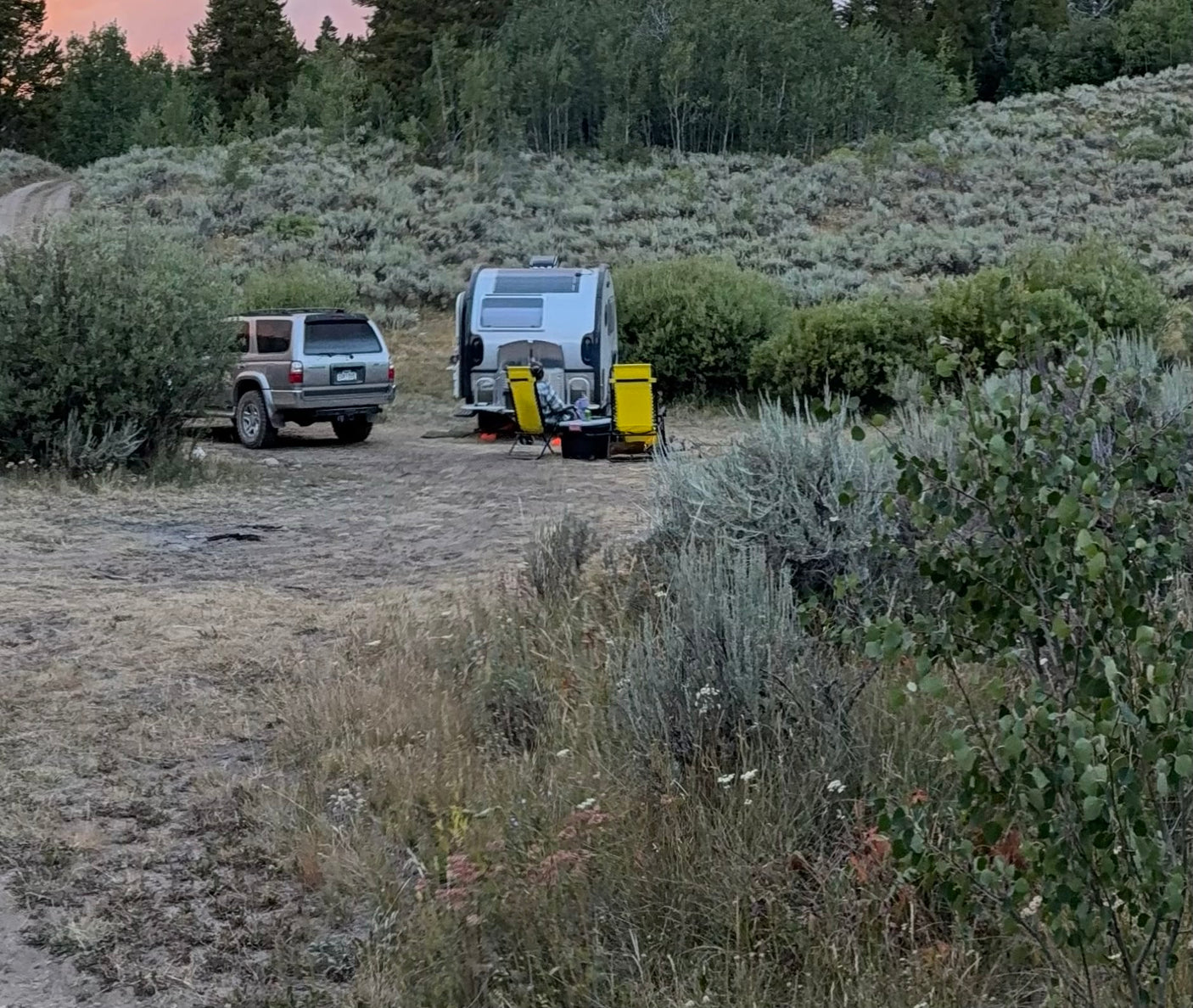An RV rental teardrop camper with yellow chairs, an SUV, and a person relaxing at a rustic campsite.