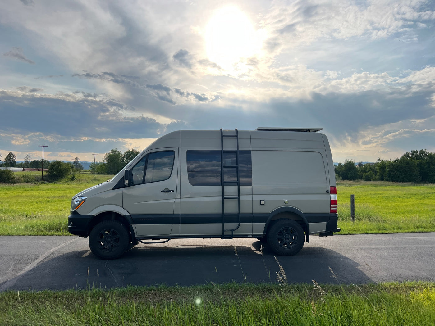 Grey camper van for RV rental on a road by lush green fields under a bright, cloudy sky with sun rays. — photo 2