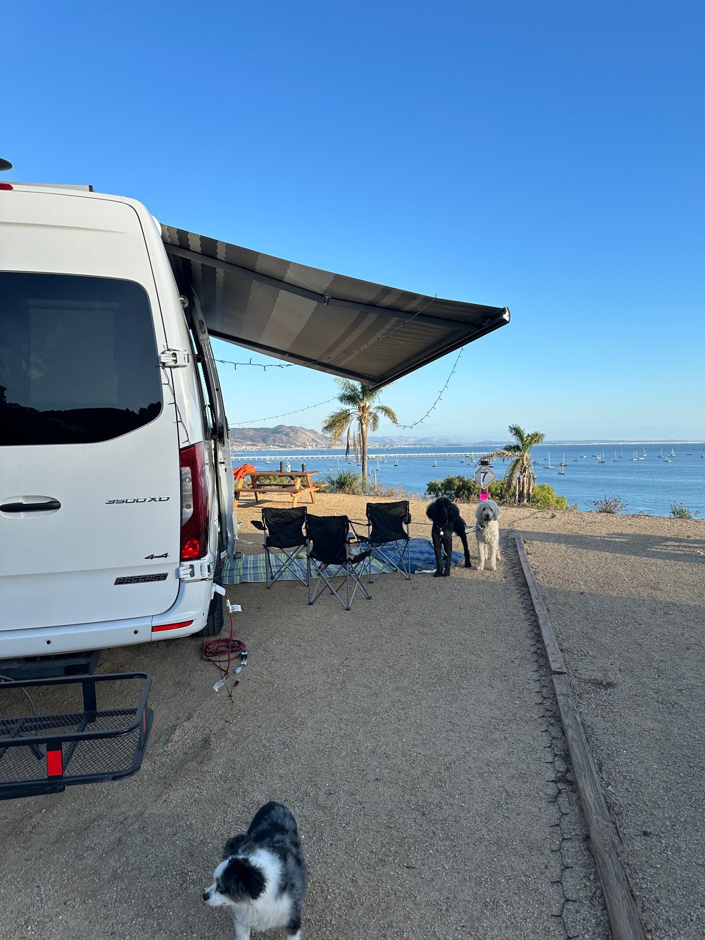 An RV rental parked by a scenic bay with boats. An extended awning, chairs, & three dogs create a peaceful campsite.