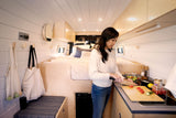 Inside a sleek RV rental, a woman chops vegetables in the compact kitchen. The comfortable sleeping area is visible behind her.