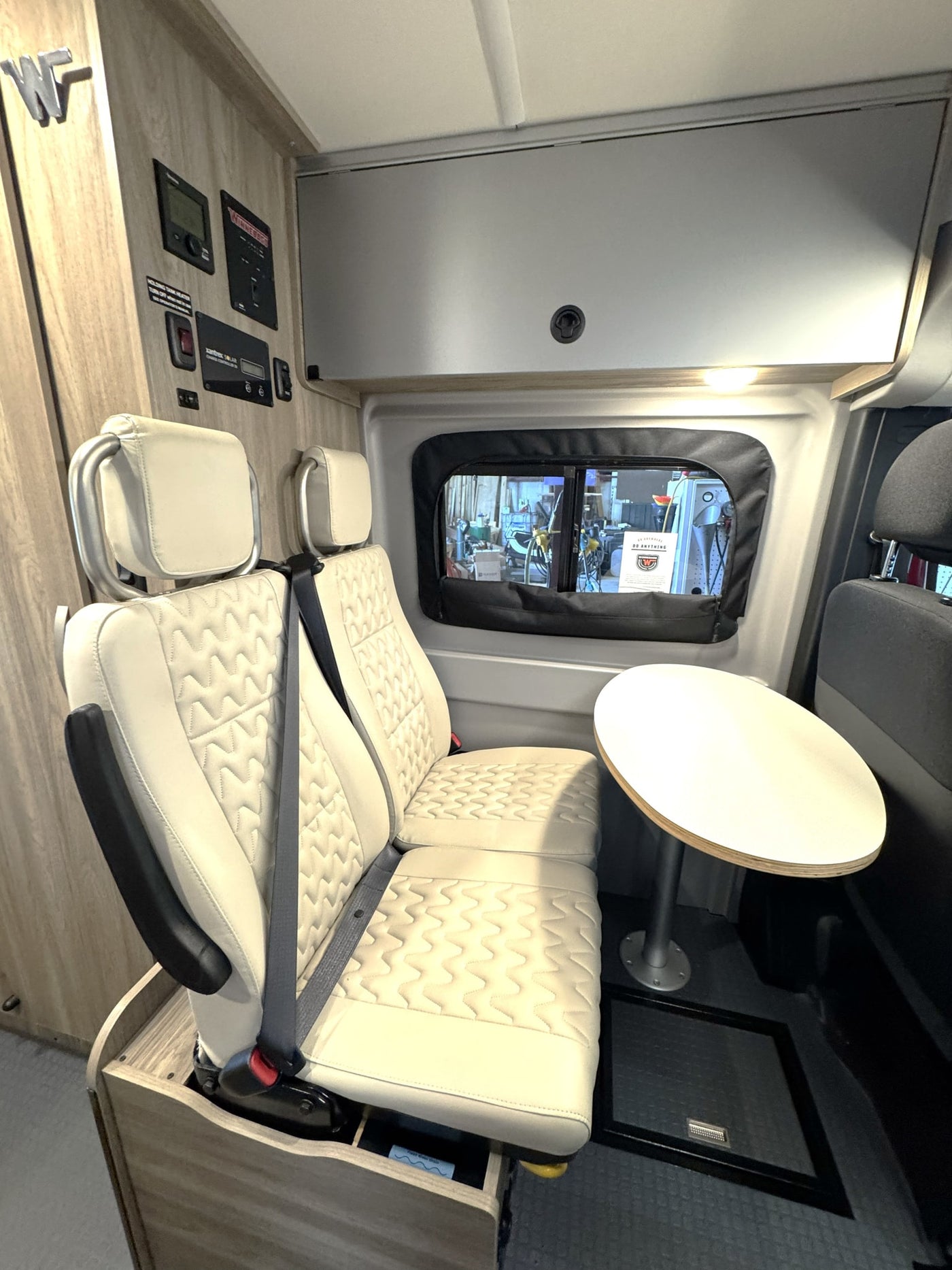 This RV rental's modern interior features cream quilted seats, a small dining table, and system controls mounted on wood-paneled walls. — photo 12