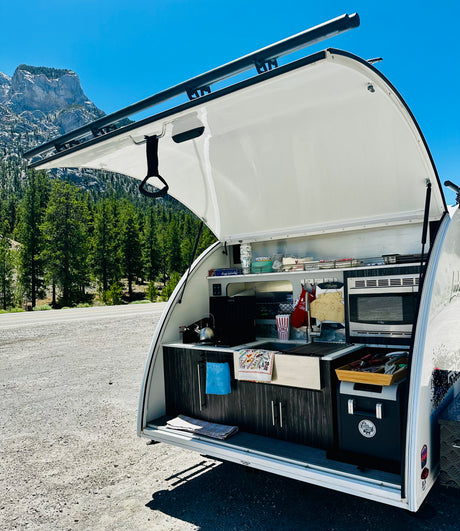 Open rear kitchen of an RV rental teardrop trailer, ready for outdoor cooking amidst mountain scenery.