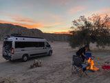 An RV rental parked in the desert at sunset. A person tends a campfire surrounded by camping chairs near a large tree.