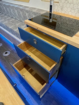 Modern RV rental kitchen featuring deep blue cabinets, open storage, induction cooktop, bench seating, and ambient blue floor lighting.