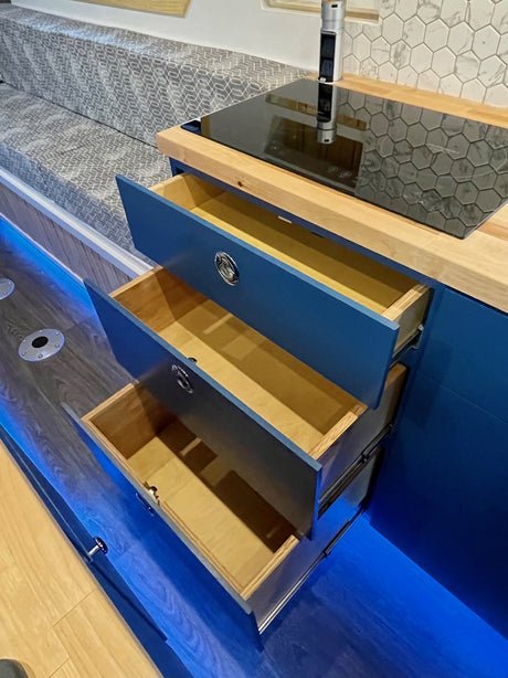 Modern RV rental kitchen featuring deep blue cabinets, open storage, induction cooktop, bench seating, and ambient blue floor lighting.