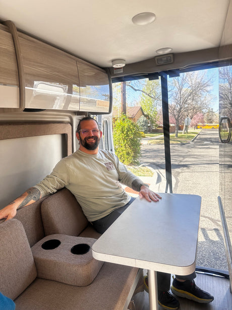 A man smiles from a cozy seat inside an RV rental, with a dining table and large window showing a sunny street.