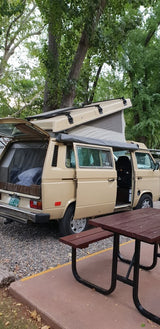 A tan pop-top camper van (RV rental) is set up for camping at a shaded site with a picnic table.