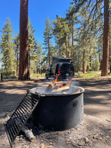 Campfire burns bright in a serene forest campground, with a black Mercedes RV rental van parked under clear blue skies.