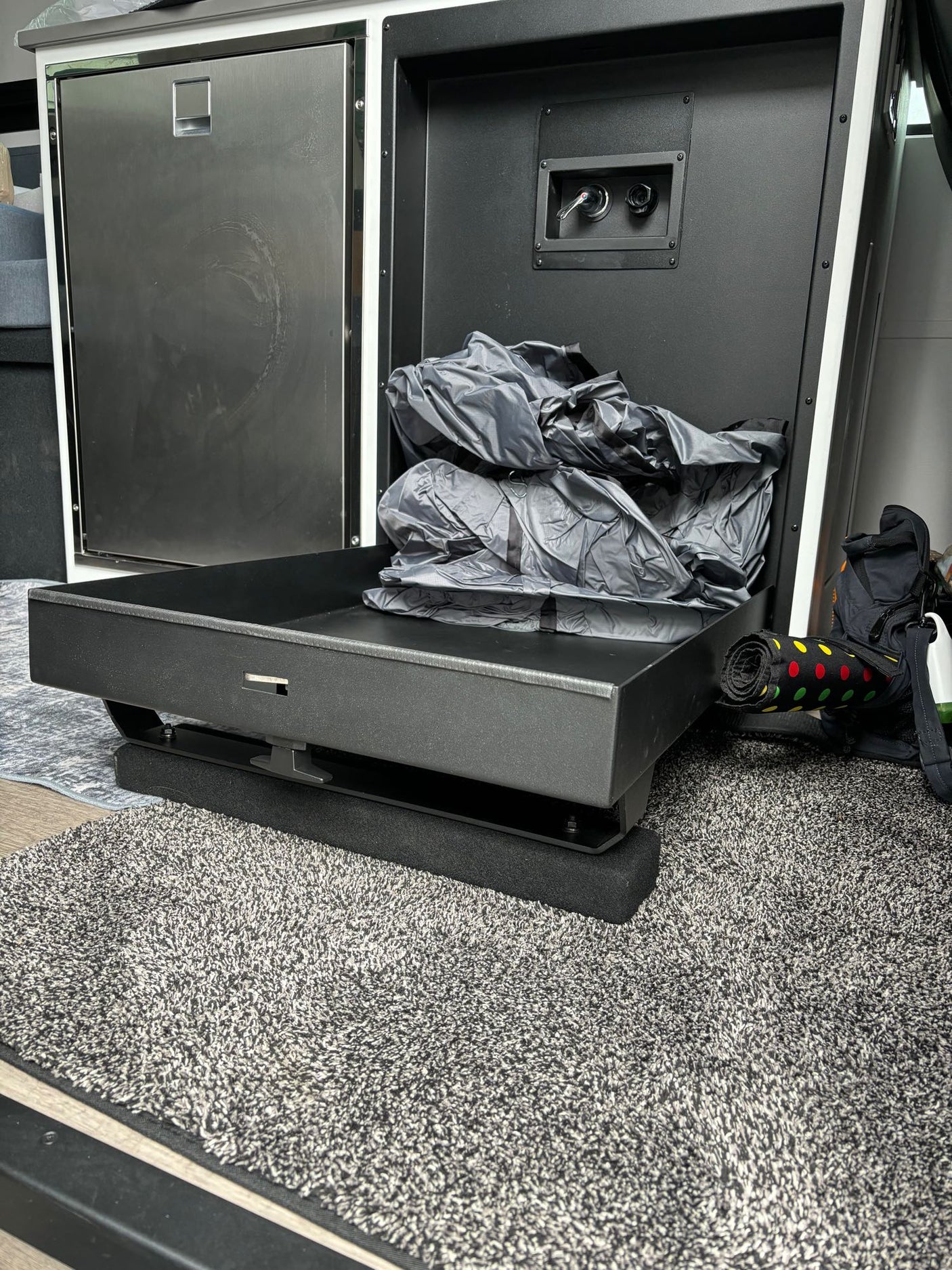 Compact RV rental interior featuring a mini-fridge, water hookups, and pull-out storage with gear on a textured floor.