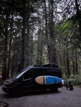 RV rental camper van in a lush, mossy forest. A paddleboard is strapped to the side; gear dries off the back.