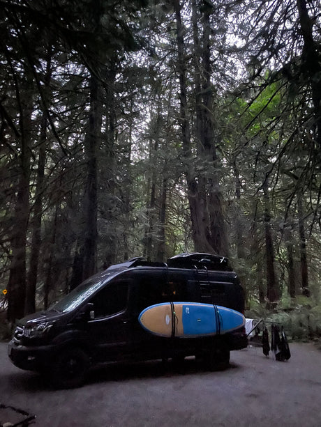 RV rental camper van in a lush, mossy forest. A paddleboard is strapped to the side; gear dries off the back.