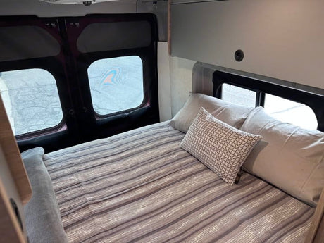 Cozy RV rental bed with striped cover and pillows, surrounded by windows in a comfortable camper interior.