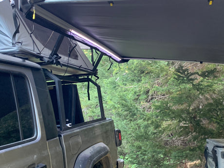 Adventure-ready RV rental: Jeep Gladiator with rooftop tent & awning setup in a lush forest for camping.