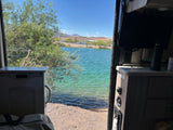 Serene lake view from an RV rental, showing clear turquoise water, a sandy shore, and mountains under a blue sky.