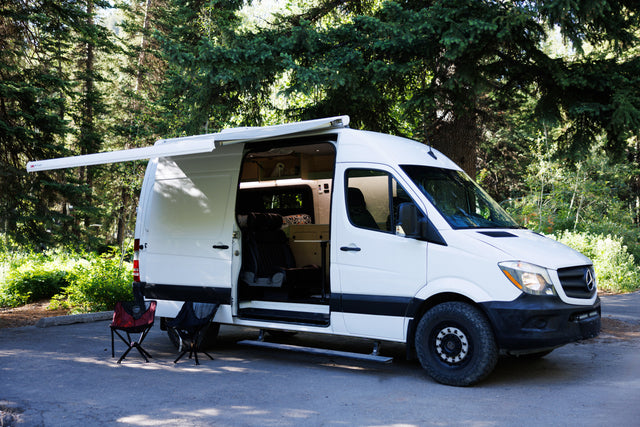 This white Mercedes Sprinter camper van, perfect for rv rental trips, is ready for camping with its side door open and awning extended.