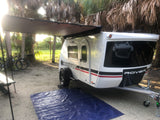 This teardrop camper, possibly an RV rental, is set up at a sunny campsite under palm trees, with an awning extended, bikes, and a tarp.