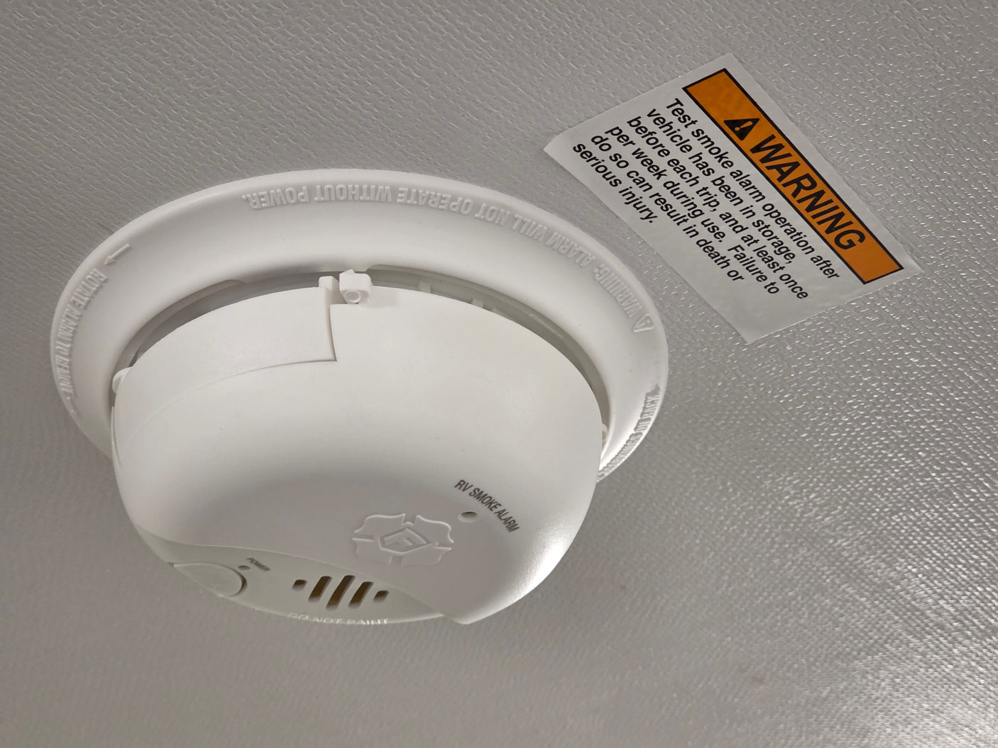 Close-up of a white RV smoke alarm and a warning sticker, emphasizing testing for safety in an RV rental.