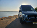 Relaxing beachside view: dark Mercedes Sprinter camper van (rv rental) on road, sand & calm water bathed in golden light.