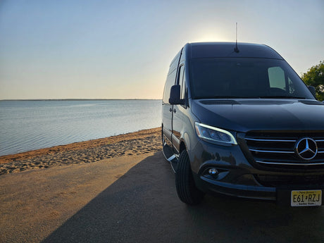 Relaxing beachside view: dark Mercedes Sprinter camper van (rv rental) on road, sand & calm water bathed in golden light.