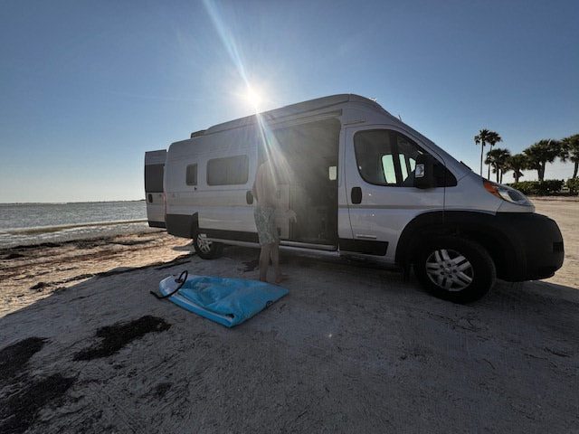 Waterfront RV rental adventure. Camper van on sandy beach by the ocean under bright sun. Person and gear ready for fun.