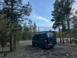 Remote camping setup: dark RV rental van, pine trees, majestic snowy mountains, and a colorful hammock ready for relaxation.