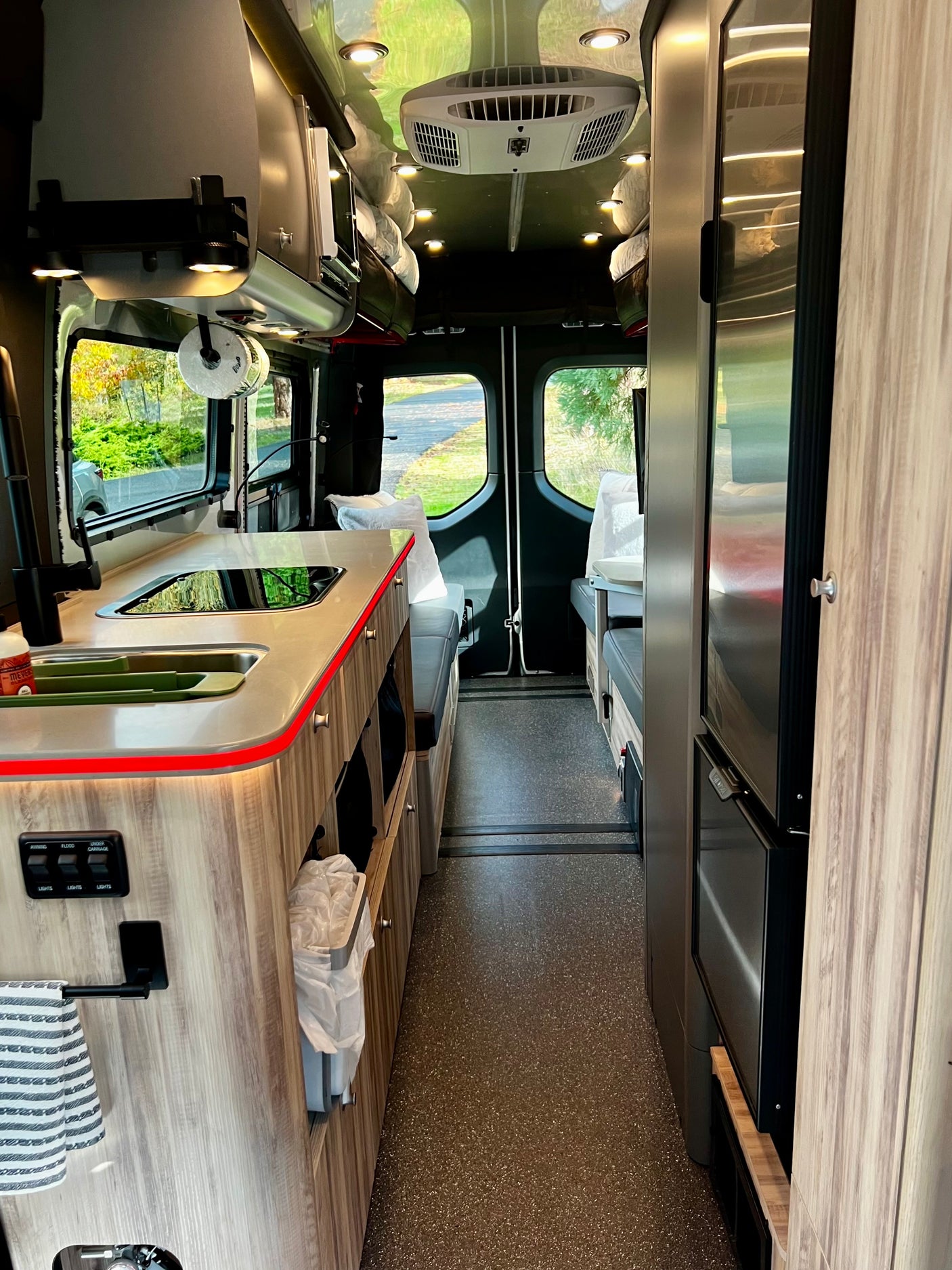 Inside a modern rv rental: narrow aisle, kitchen, fridge, and seating leading to rear windows, featuring bright overhead lighting.