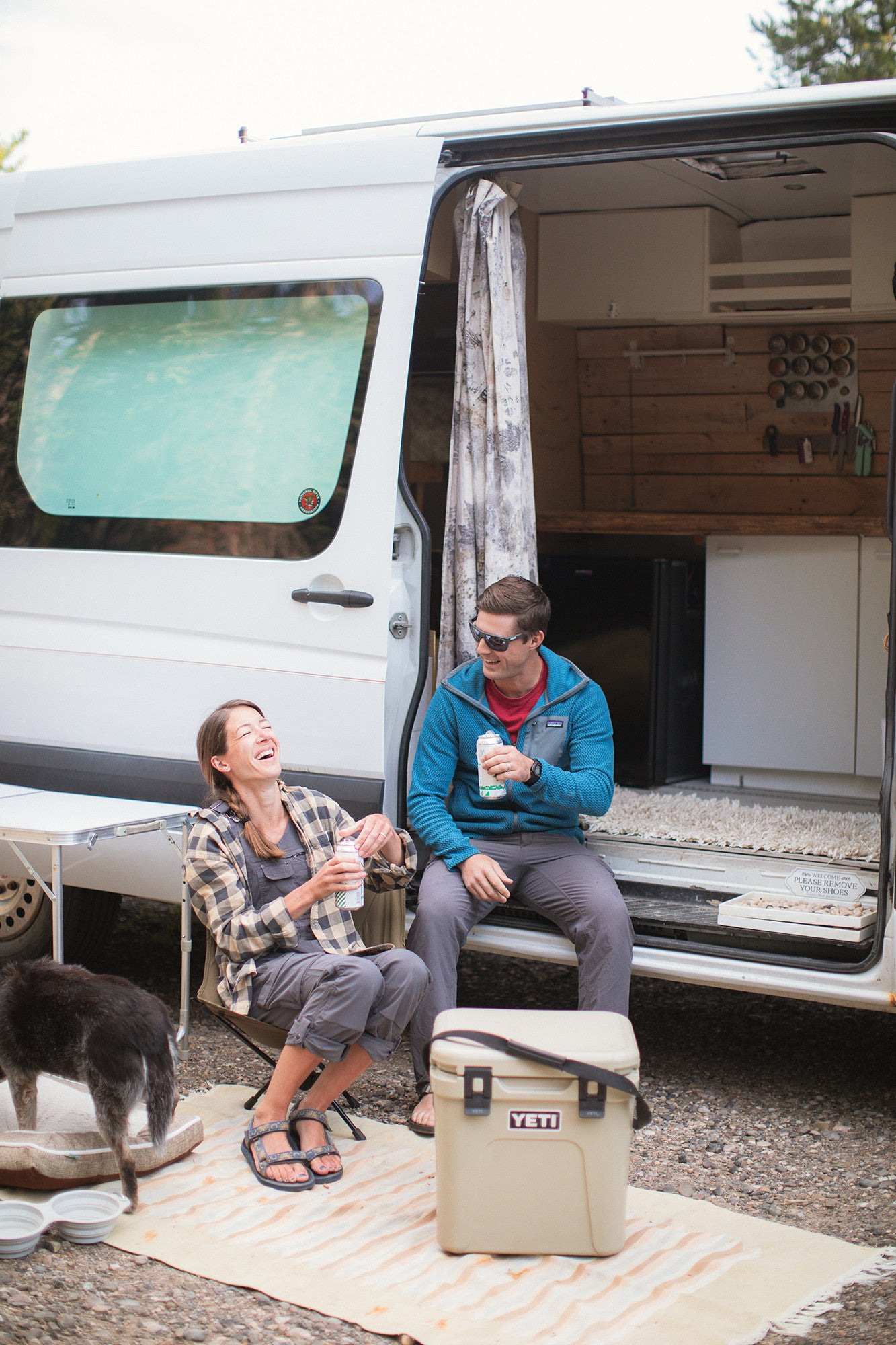 Smiling couple relaxes by their custom RV rental, enjoying van life on an adventure with their dog.