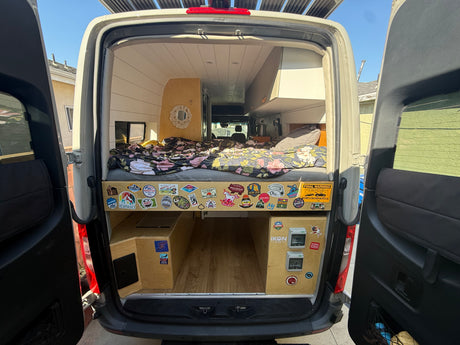 Custom camper van interior, possibly an RV rental, showing a cozy bed, under-bed storage, and control panels at the rear.